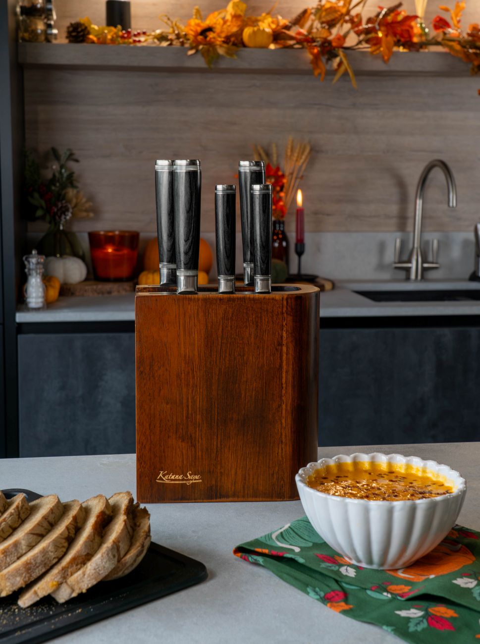 A kitchen counter with autumnal decorations, a pakkawoord Katana Saya block set and a bowl of soup with a sliced loaf of bread.