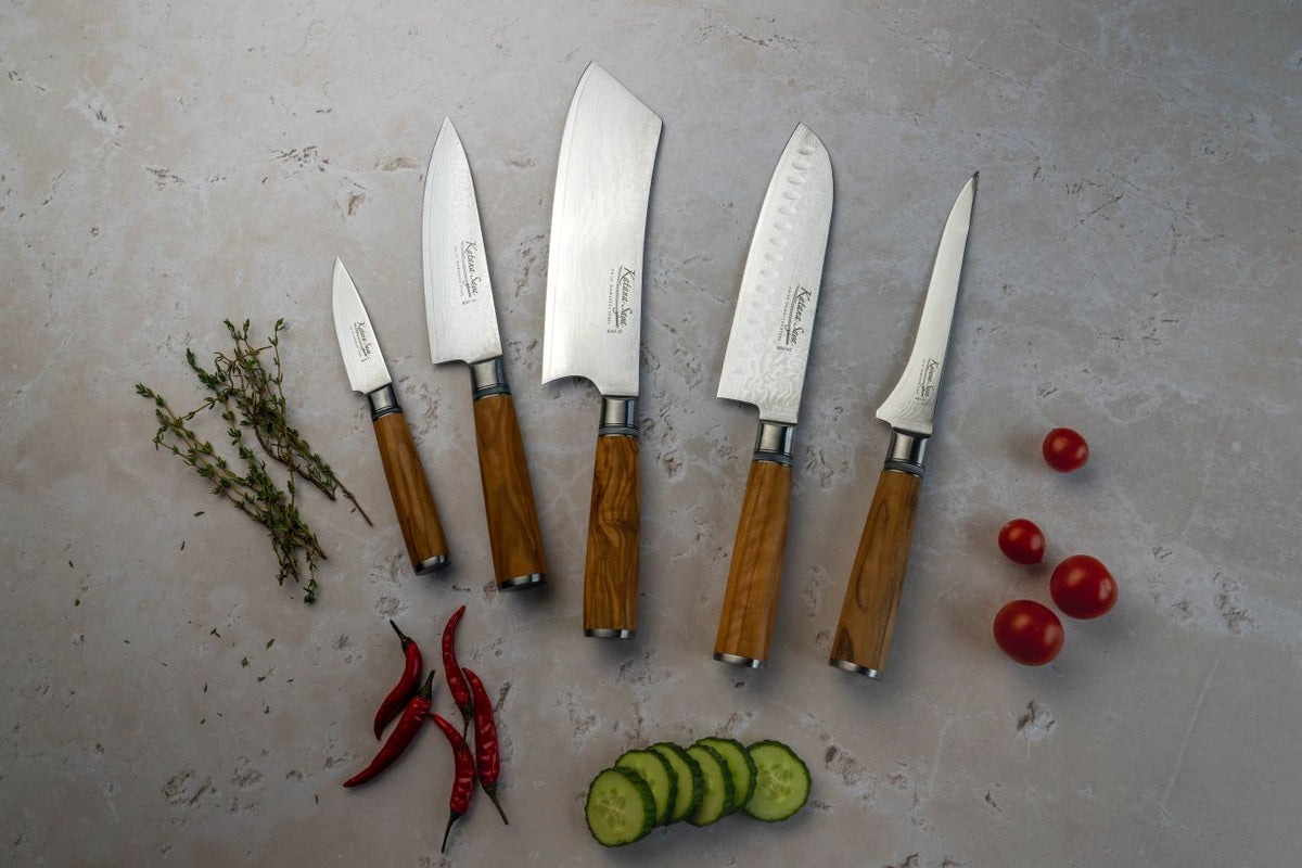 Five olivewood knives laid on a kitchen work top with chopped vegetables.