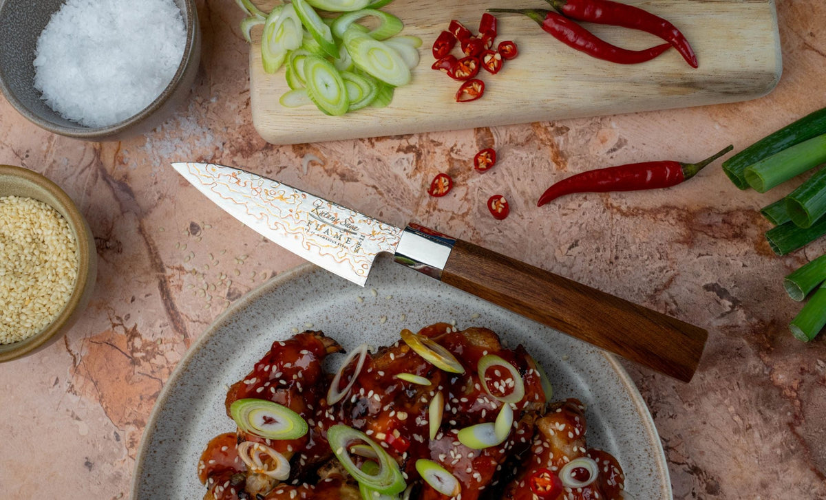 A high-end kitchen knife, the Rosewood Flame serrated brad knife, with a Damascus blade and a rose wood handle, displayed diagonally alongside its wooden sheath on a rustic wooden cutting board with a loaf of bread, rosemary, and a bowl of coarse salt.