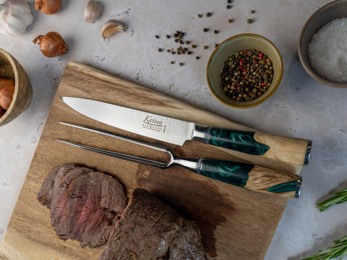 The Katana Elements Rainforest kitchen carving set with a knife and fork resting on a wooden cutting board beside sliced roast meat, surrounded by garlic, peppercorns, salt, and herbs on a light stone surface.