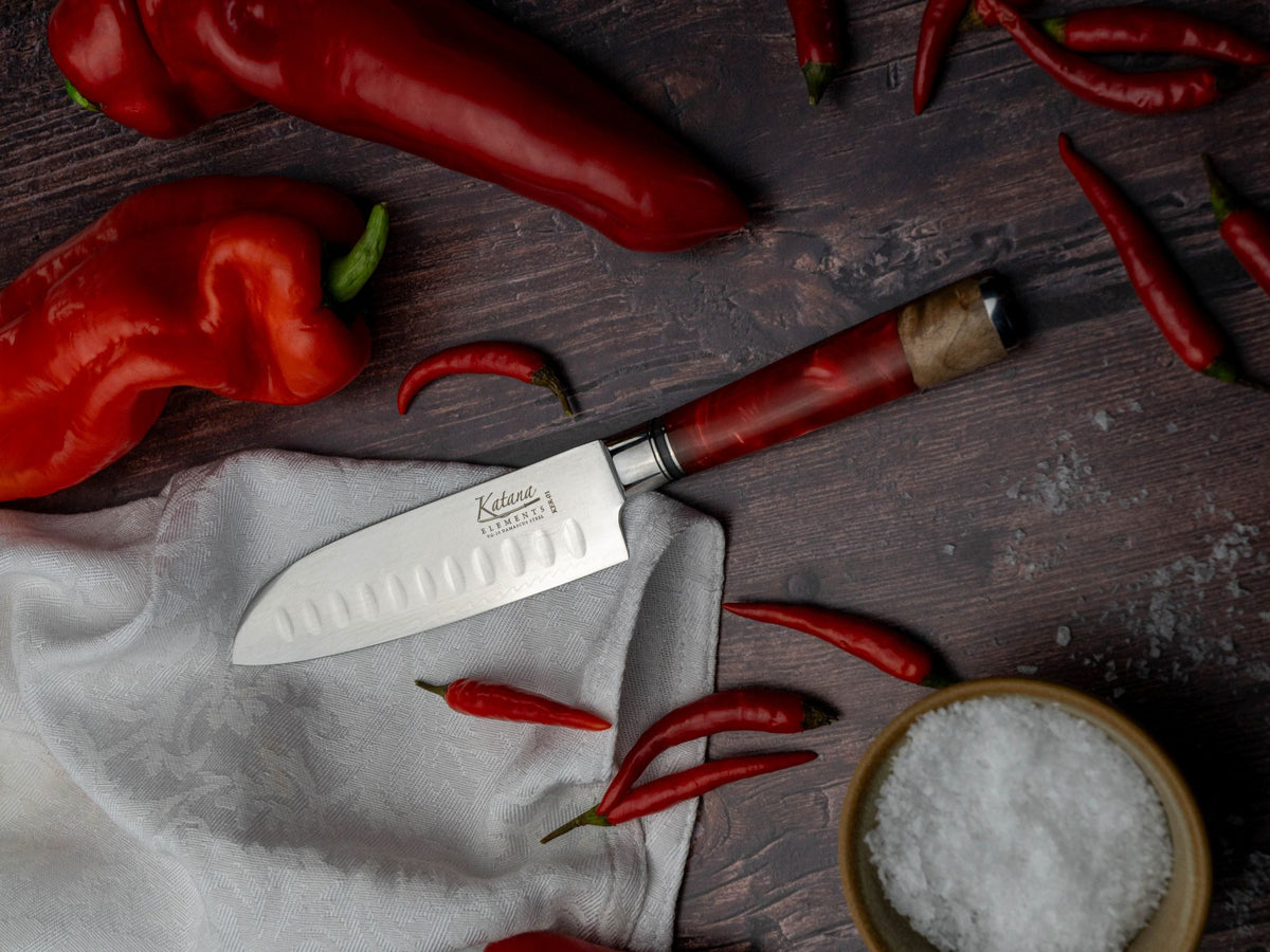 A Katana Elements Volcanic fluted santoku kitchen knife with a silver blade and reddish-brown wooden handle lies on a white cloth over a dark wooden surface, surrounded by red chili peppers and a small bowl of coarse salt.