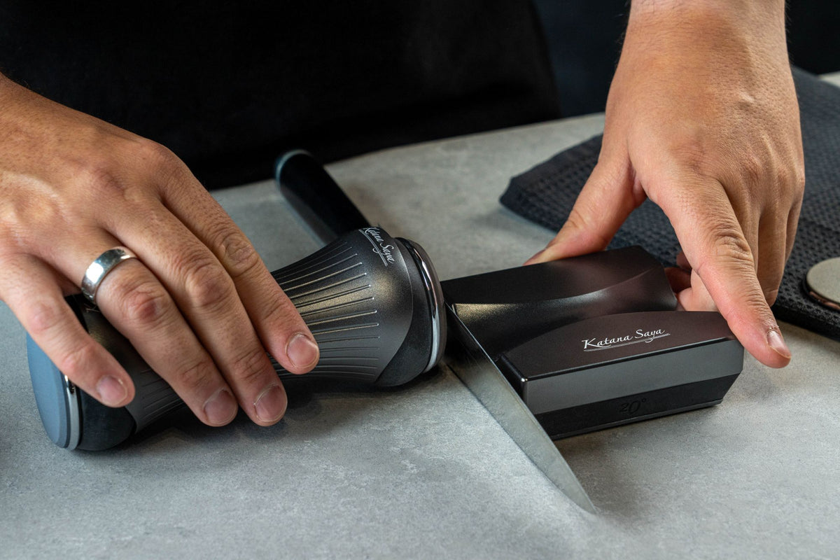 A close-up of a person's hands holding the Katana Saya Roller Sharpener to sharpen a Pakkawood kitchen knife on a light gray surface.