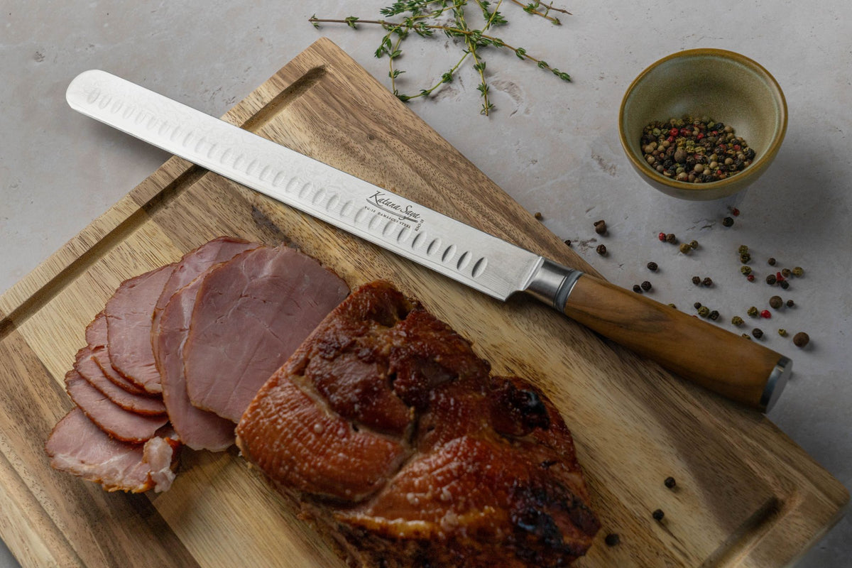 Katana Saya Olive Wood Slicing Knife with Damascus steel fluted blade on wooden chopping board next to sliced gammon, pepper corns and thyme.