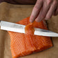 A close-up of a Pakkawood sashimi knife being used to slice a thick salmon fillet on a sheet of brown parchment paper, with a person's hands visible holding the knife and guiding the salmon.