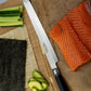 A Katana Saya Pakkwood sashimi kitchen knife with a black wooden handle and Damscus steel single bevel blade lies on a brown cutting board next to a salmon fillet. In the background, another cutting board holds nori seaweed, sliced avocado, celery, and cucumber on a bamboo mat.