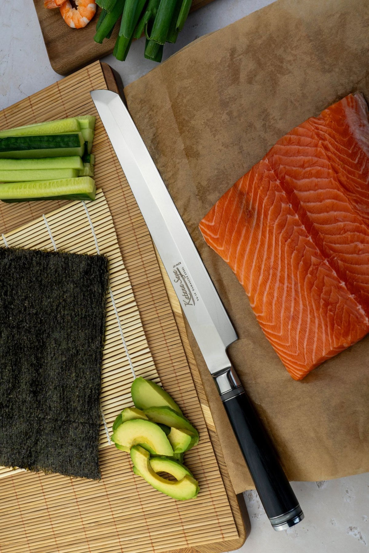 A Katana Saya Pakkwood sashimi kitchen knife with a black wooden handle and Damscus steel single bevel blade lies on a brown cutting board next to a salmon fillet. In the background, another cutting board holds nori seaweed, sliced avocado, celery, and cucumber on a bamboo mat.