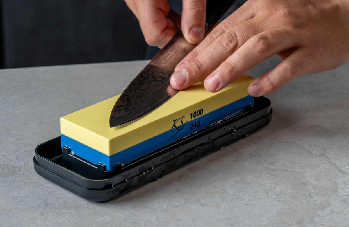 A close-up of a person sharpening a Katana Saya knife on a dual-sided sharpening stone. The whetstone has a yellow top side marked with '1000' grit and '280' grit on the blue bottom side, resting in a black plastic holder on a gray countertop.