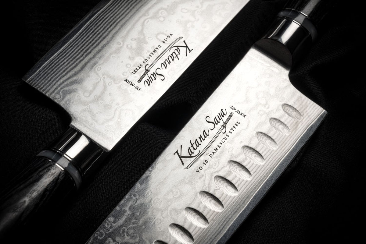 Two Katana Saya Damascus steel kitchen knives with textured patterned blades and black handles, displayed diagonally on a dark background.